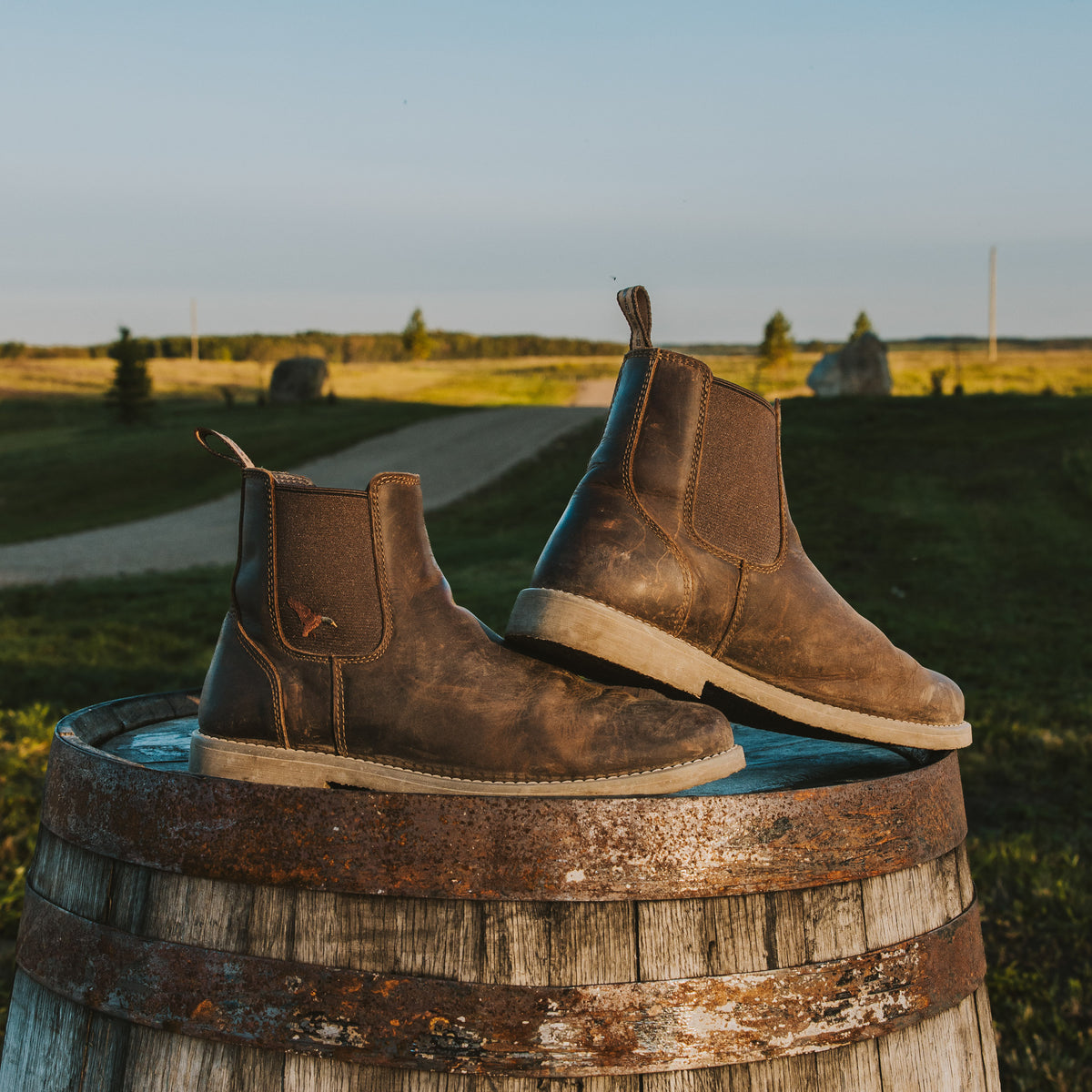 A pair of Dive Bomb Dark Walnut Heritage Boot with distressed detailing, resting on top of a weathered wooden barrel in an outdoor rural setting with a grassy field and path in the background.