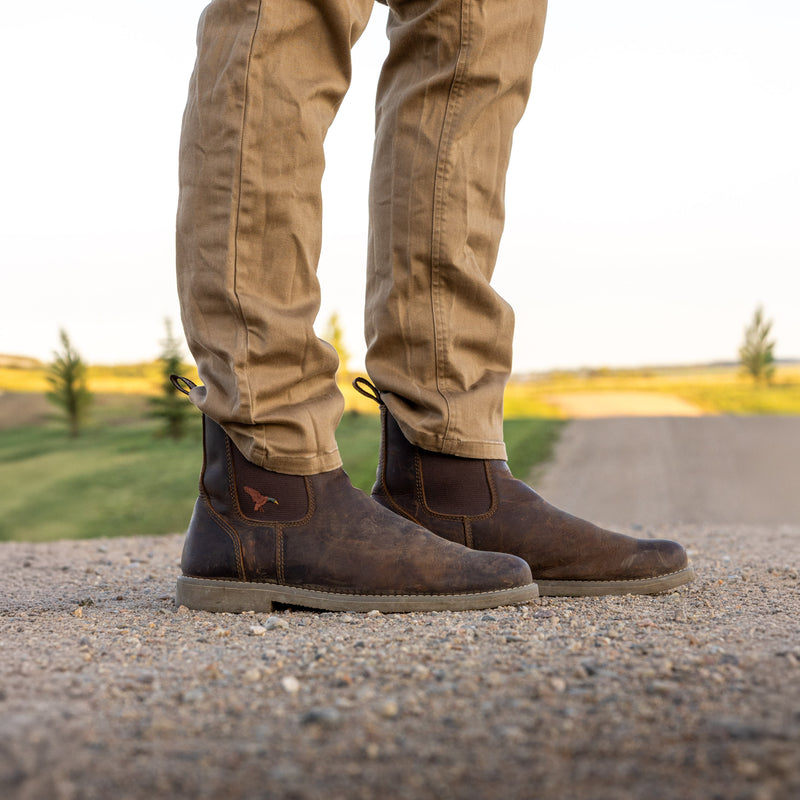A person wearing Dive Bomb Dark Walnut Heritage Boot and tan pants, standing on a gravel path outdoors with a blurred field in the background.