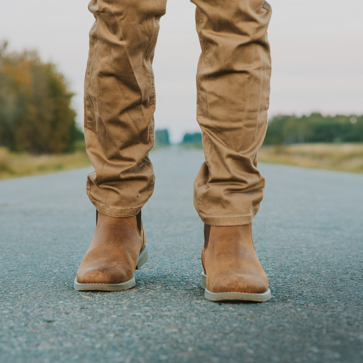 A person standing on an asphalt road wearing Dive Bomb Tobacco Brown Heritage Boot, a Chelsea style boot, with trees blurred in the background.