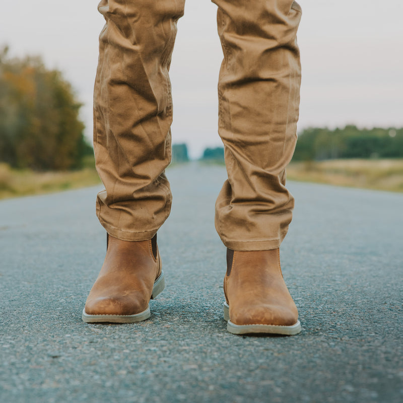 A person standing on an asphalt road wearing Dive Bomb Tobacco Brown Heritage Boot, a Chelsea style boot, with trees blurred in the background.