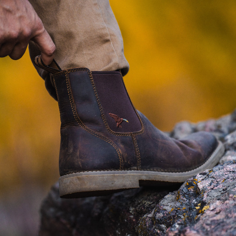 A close-up of a person wearing Dive Bomb Dark Walnut Heritage Boot with tan pants, standing on a rock with a blurred yellow autumn background.