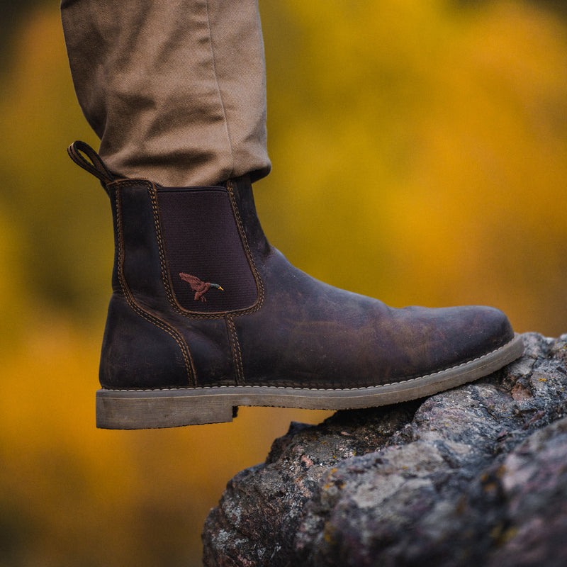 Dive Bomb Dark Walnut Heritage Boot Chelsea-style boot with a small mallard logo on the side, worn with tan pants and resting on a rock.