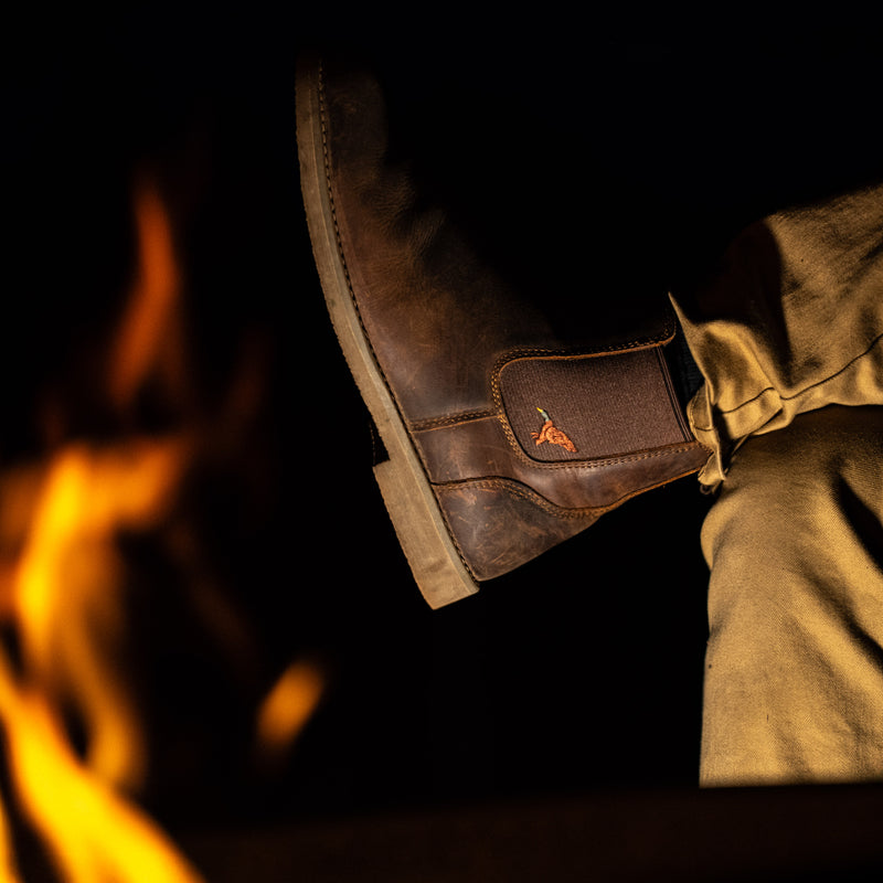 A close-up of a person's leg wearing a Dive Bomb Dark Walnut Heritage Boot in a Chelsea style with tan trousers, set against a dark background with warm orange fire flames on the left.