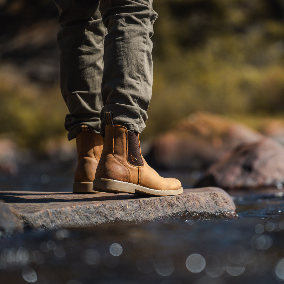 A person standing on a rock in shallow water wearing Dive Bomb Tobacco Brown Heritage Boot and olive pants, with a blurred outdoor background of rocks and greenery.
