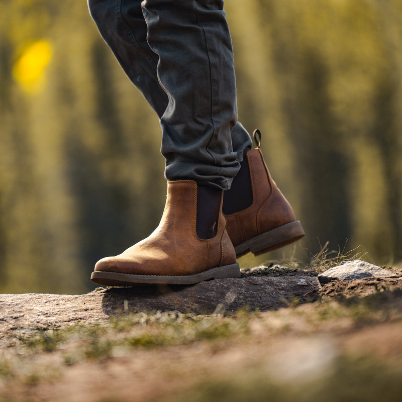 A person wearing dark pants and Dive Bomb Tobacco Brown Heritage Boot, standing on a log in an outdoor forest setting.