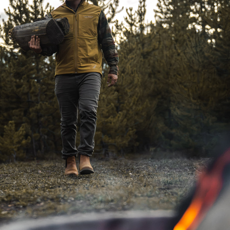 A person walking outdoors in a forest while wearing a tan zip-front vest over a camouflage long-sleeve shirt, dark gray pants, and Dive Bomb Tobacco Brown Heritage Boot, with a log in one hand.