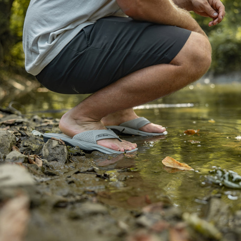 Grey Flights sandal pair worn crouched at a rocky creek edge, feet in the shallow water