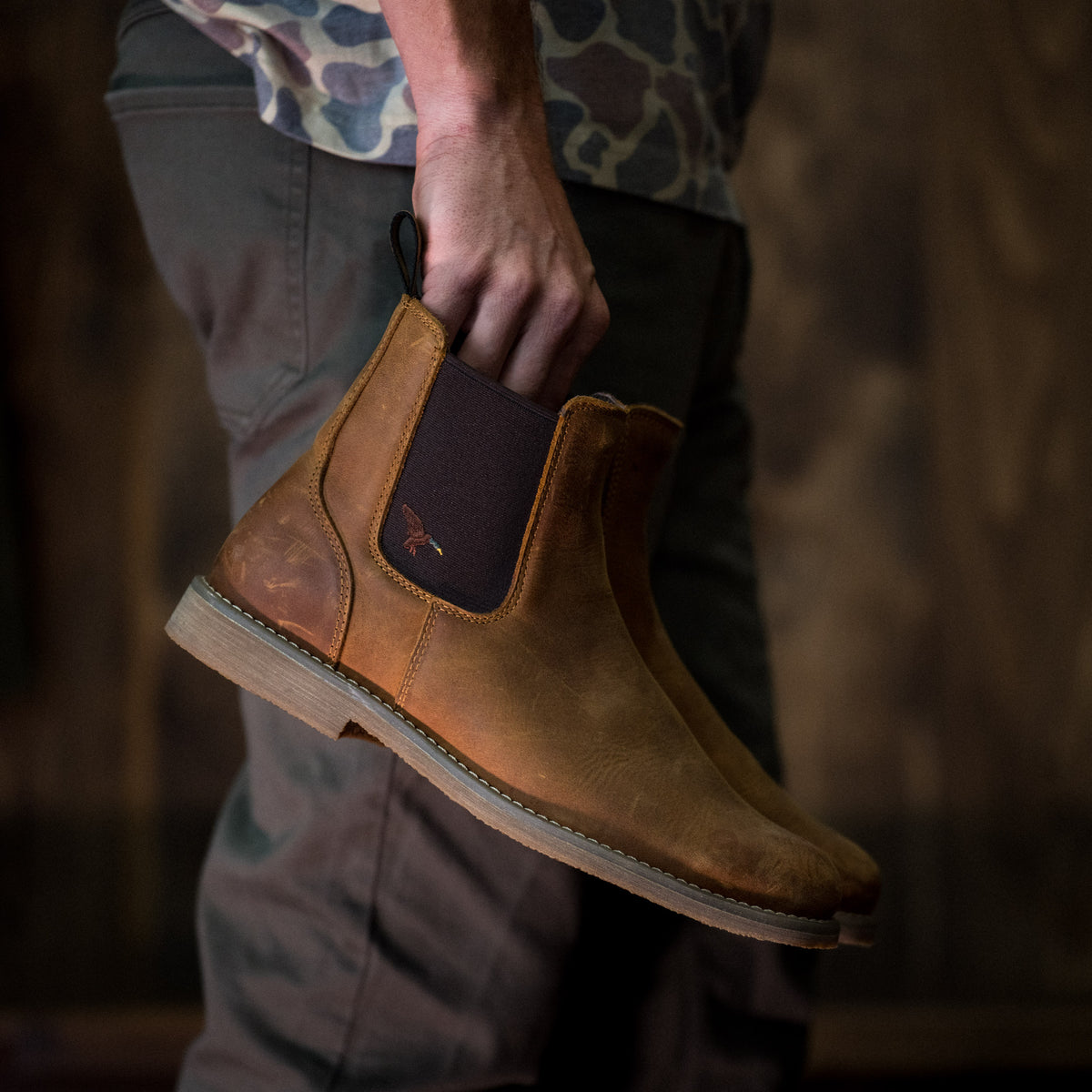 A person holds up Dive Bomb Tobacco Brown Heritage Boot against a dark rustic background. The boot has a dark brown elastic side panel and a light-colored sole.